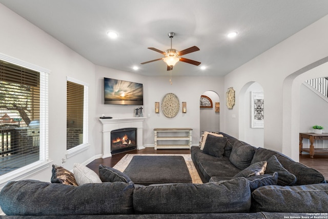 living area with arched walkways, a warm lit fireplace, dark wood finished floors, and a ceiling fan