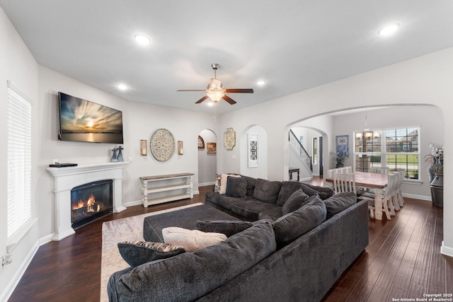 living area featuring arched walkways, a glass covered fireplace, ceiling fan with notable chandelier, and dark wood-style flooring