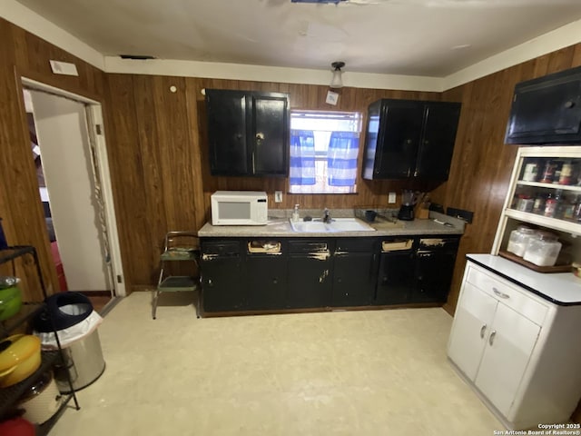 kitchen with white microwave, wood walls, light countertops, dark cabinetry, and a sink