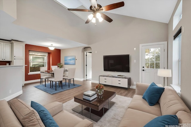 living room with baseboards, ceiling fan, light wood-type flooring, a towering ceiling, and arched walkways