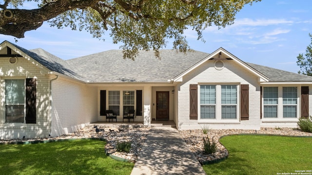 ranch-style home featuring brick siding, covered porch, a front lawn, and roof with shingles