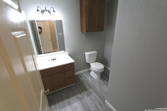 bathroom with toilet, wood finished floors, baseboards, vanity, and a textured wall