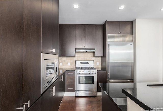 kitchen featuring under cabinet range hood, backsplash, dark brown cabinetry, built in appliances, and dark wood-style flooring