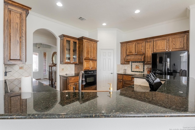 kitchen featuring dark stone countertops, arched walkways, black appliances, glass insert cabinets, and brown cabinets