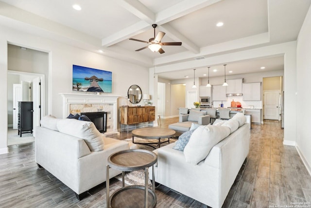 living area with wood finish floors, beam ceiling, a stone fireplace, and a ceiling fan