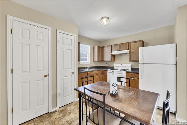 kitchen with under cabinet range hood, brown cabinetry, white appliances, and dark countertops