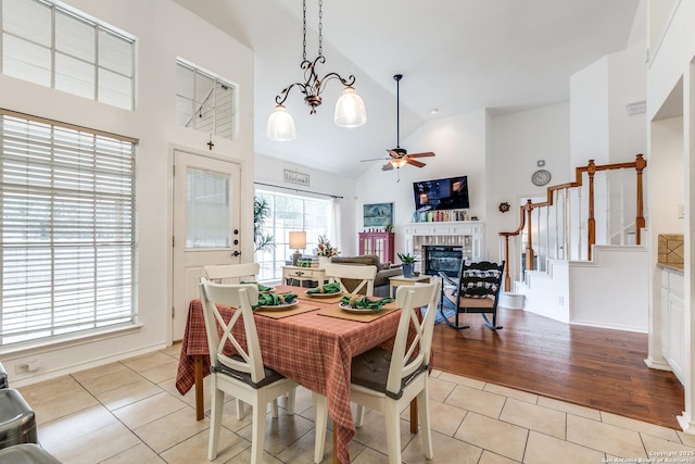 dining room featuring a brick fireplace, stairway, ceiling fan with notable chandelier, light tile patterned flooring, and high vaulted ceiling