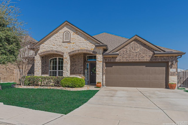 french country home with concrete driveway, a front yard, roof with shingles, stone siding, and an attached garage