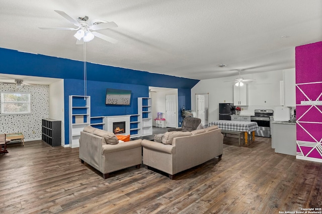 living room with dark wood-style floors, wallpapered walls, a lit fireplace, ceiling fan, and a textured ceiling