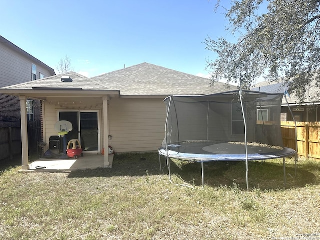 back of house featuring a shingled roof, a trampoline, fence, a lawn, and a patio area