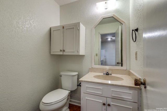 half bathroom with vanity, toilet, a textured wall, and baseboards