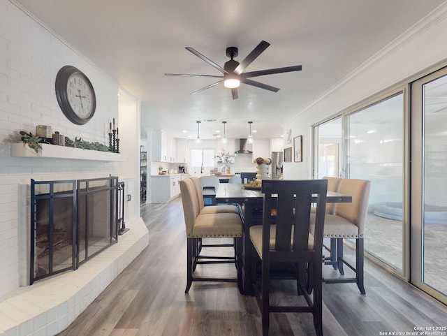 dining area featuring wood finished floors, a brick fireplace, a ceiling fan, and ornamental molding