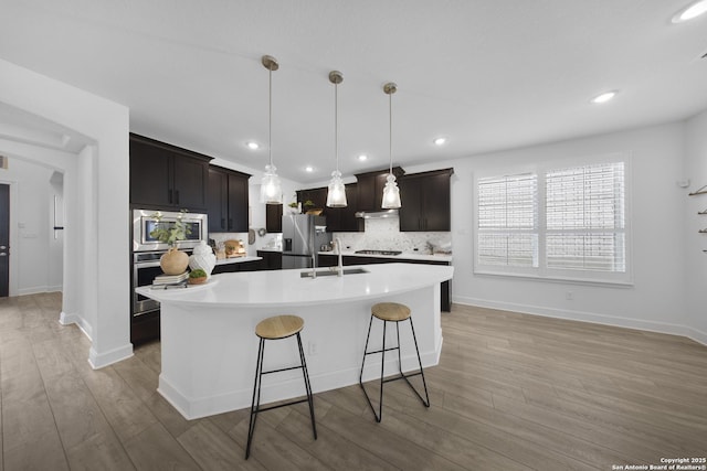 kitchen featuring light wood-type flooring, an island with sink, backsplash, stainless steel appliances, and light countertops