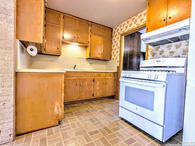 kitchen with wallpapered walls, brick floor, light countertops, under cabinet range hood, and white gas range