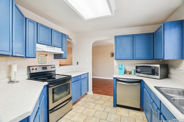 kitchen with light countertops, blue cabinetry, under cabinet range hood, appliances with stainless steel finishes, and arched walkways