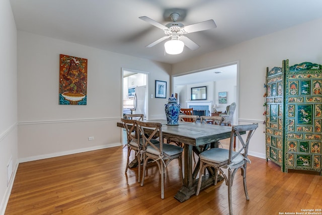 dining space with a fireplace, baseboards, light wood-type flooring, and ceiling fan