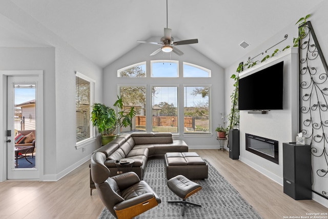 living room with visible vents, a ceiling fan, a glass covered fireplace, light wood finished floors, and vaulted ceiling