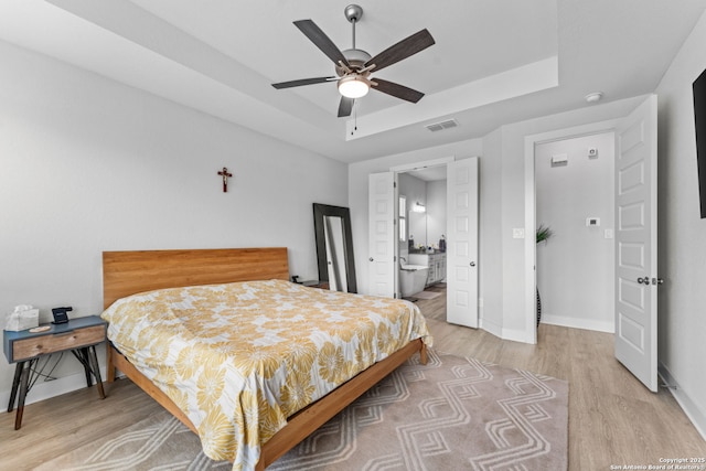 bedroom featuring visible vents, baseboards, a tray ceiling, light wood-style floors, and connected bathroom