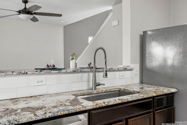 kitchen featuring stainless steel dishwasher, light stone counters, dark brown cabinets, a sink, and a ceiling fan
