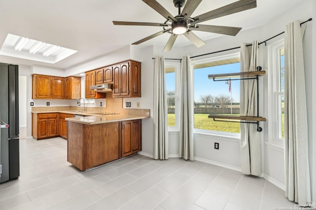 kitchen featuring a ceiling fan, under cabinet range hood, a peninsula, brown cabinetry, and light countertops