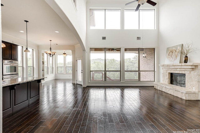 unfurnished living room with ceiling fan with notable chandelier, dark wood finished floors, a stone fireplace, and a wealth of natural light