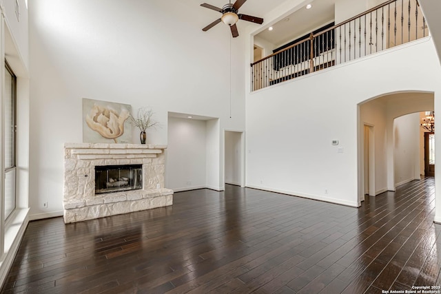 unfurnished living room with arched walkways, a stone fireplace, ceiling fan with notable chandelier, baseboards, and a towering ceiling