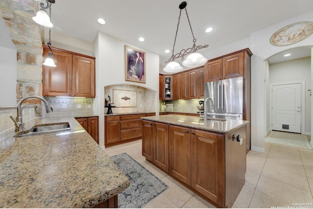 kitchen featuring light stone countertops, a center island with sink, a sink, brown cabinetry, and freestanding refrigerator