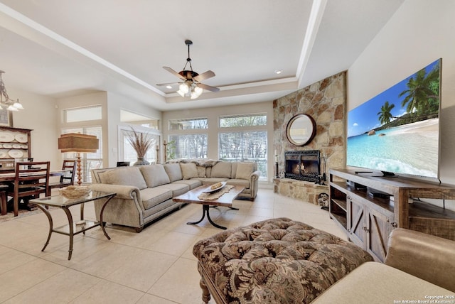 living room featuring light tile patterned flooring, a tray ceiling, a stone fireplace, ceiling fan, and recessed lighting