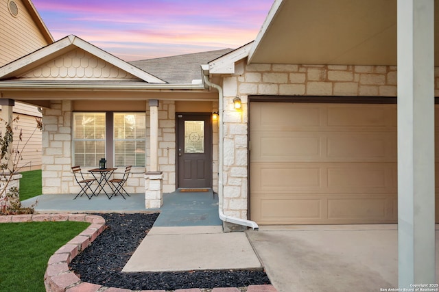 property entrance with stone siding, an attached garage, a shingled roof, and covered porch