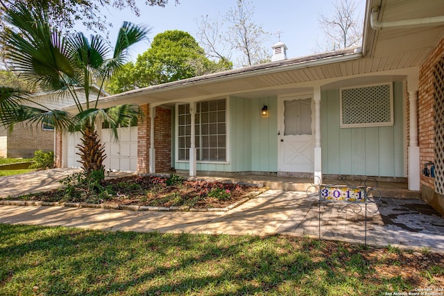 doorway to property featuring driveway, a chimney, a porch, brick siding, and a garage