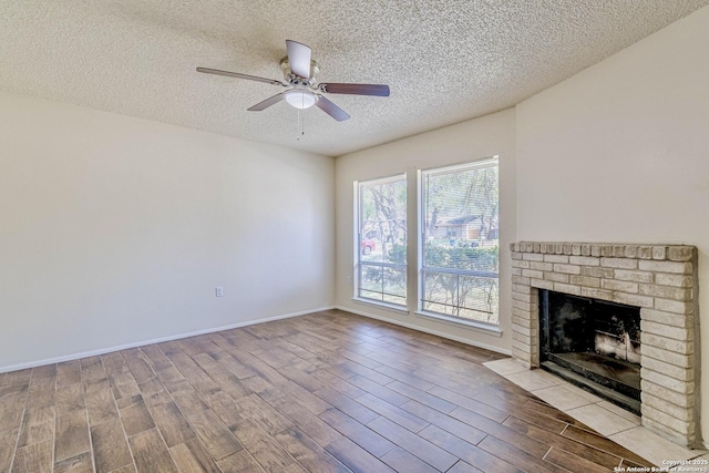 unfurnished living room featuring wood finished floors, a textured ceiling, a brick fireplace, baseboards, and ceiling fan