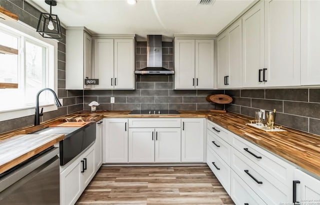 kitchen featuring black electric cooktop, butcher block countertops, stainless steel dishwasher, a sink, and wall chimney range hood