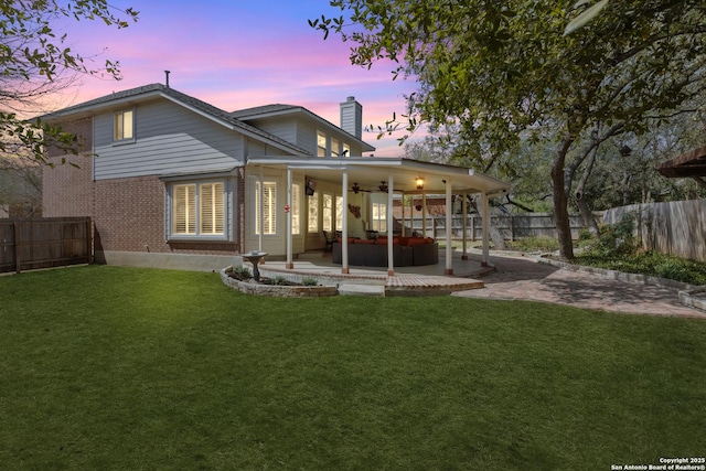 back of property at dusk featuring a patio, a fenced backyard, a ceiling fan, brick siding, and a lawn