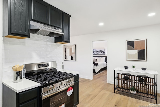 kitchen featuring stainless steel gas range oven, light wood-type flooring, under cabinet range hood, backsplash, and dark cabinets