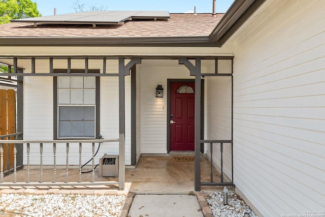 doorway to property featuring solar panels, a shingled roof, and a porch