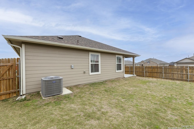 rear view of property featuring cooling unit, a shingled roof, fence, and a lawn
