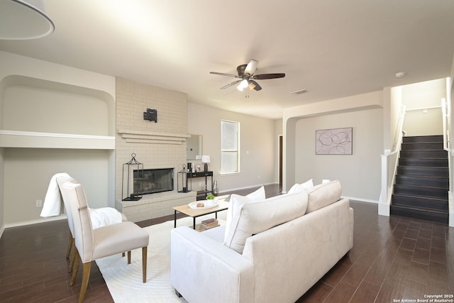 living room featuring stairway, a ceiling fan, a brick fireplace, dark wood-style floors, and baseboards