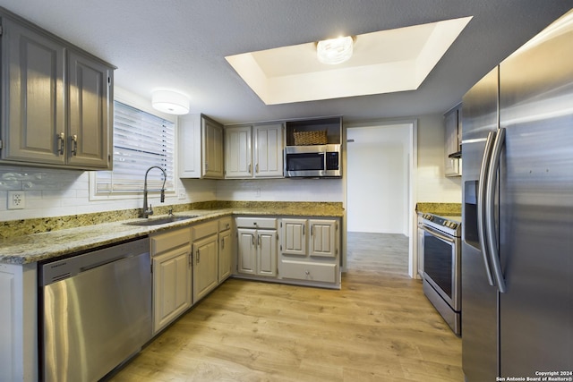 kitchen featuring light wood-type flooring, stainless steel appliances, a raised ceiling, a sink, and light stone countertops