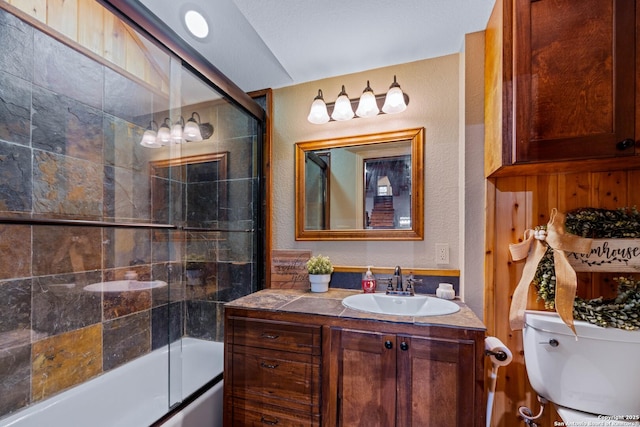 full bath with a textured wall, vanity, shower / bath combination with glass door, and toilet