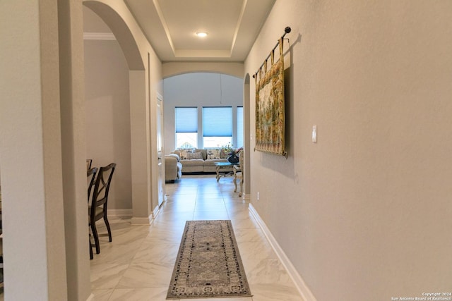 corridor with arched walkways, marble finish floor, a tray ceiling, and baseboards