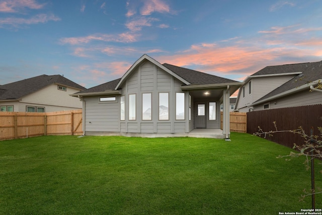 back of property at dusk with roof with shingles, a fenced backyard, and a lawn