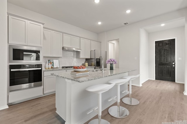 kitchen featuring under cabinet range hood, light stone countertops, visible vents, a center island with sink, and stainless steel appliances
