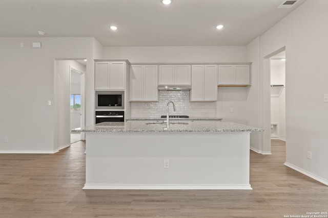 kitchen featuring oven, under cabinet range hood, a center island with sink, backsplash, and built in microwave