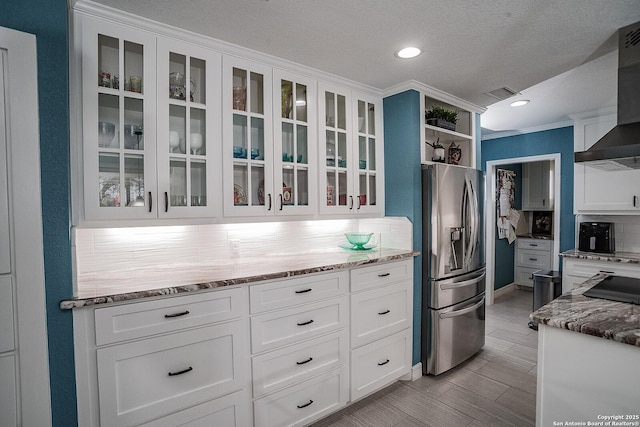 kitchen with white cabinets, visible vents, stainless steel refrigerator with ice dispenser, light stone counters, and wall chimney range hood