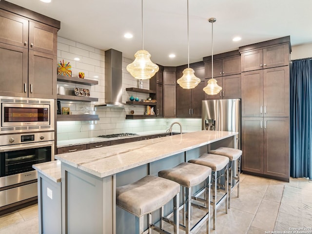 kitchen featuring open shelves, decorative backsplash, wall chimney range hood, a kitchen island with sink, and appliances with stainless steel finishes