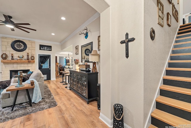 living room featuring stairs, wood finished floors, a stone fireplace, ceiling fan, and ornamental molding