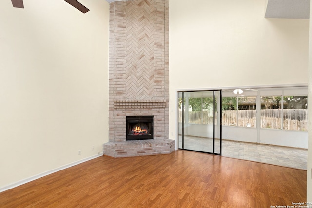 unfurnished living room featuring wood finished floors, a towering ceiling, a brick fireplace, and ceiling fan