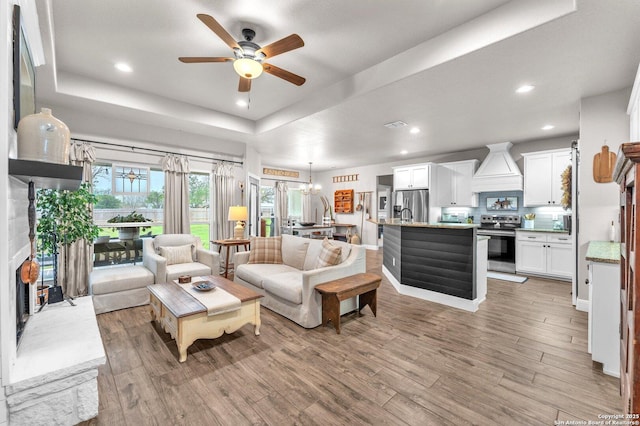 living area featuring light wood-type flooring, baseboards, recessed lighting, a tray ceiling, and ceiling fan with notable chandelier