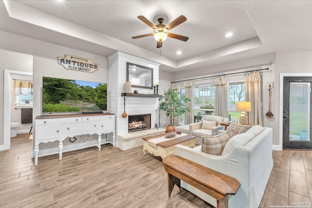 living area featuring baseboards, a ceiling fan, a tray ceiling, a large fireplace, and light wood-style flooring