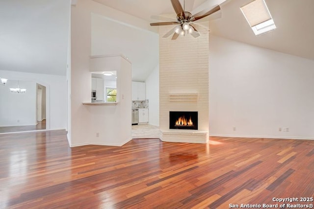 unfurnished living room with wood finished floors, a skylight, a brick fireplace, baseboards, and ceiling fan with notable chandelier